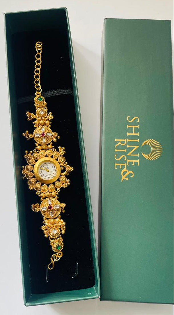 An ornate gold-toned wrist watch with a round dial and decorative jewel-like embellishments, displayed inside an open dark green presentation box next to its matching green box lid. The lid is printed with the brand name 'SHINE RISE'.