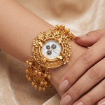 A close-up of a woman's wrist and hand wearing an ornate gold watch with a decorative leaf-patterned bezel, dangling gold charms, and a white dial.