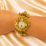 An ornate gold-tone watch with a wide, sculptural bracelet featuring floral and leaf motifs, worn on a wrist against a draped champagne satin background.