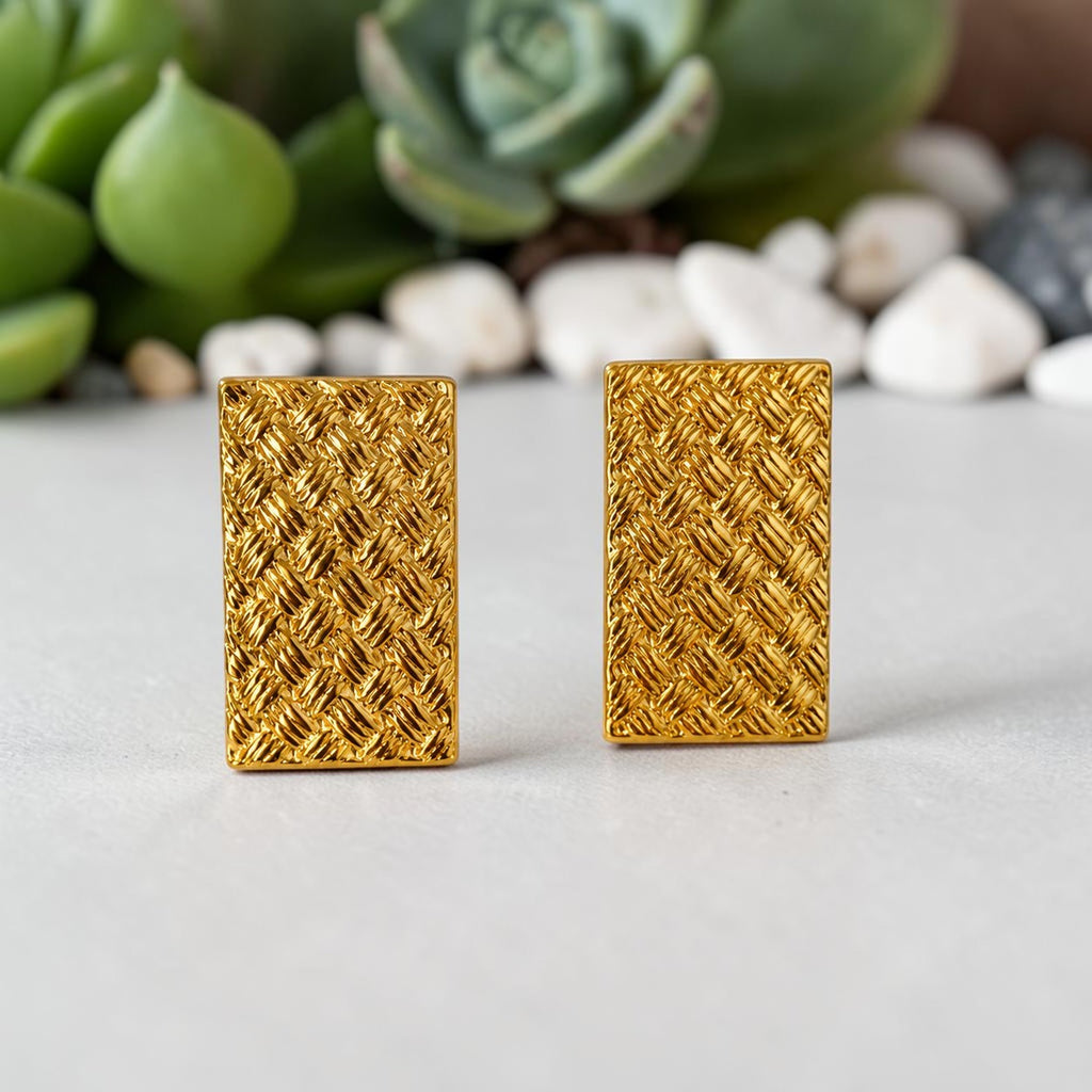 Pair of golden rectangular earrings on a light surface with a blurred natural background