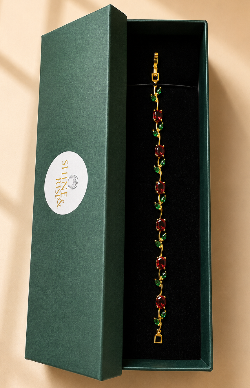 A gold-tone chain bracelet with red and green floral details, presented inside an open dark green jewelry box on a warm beige background.
