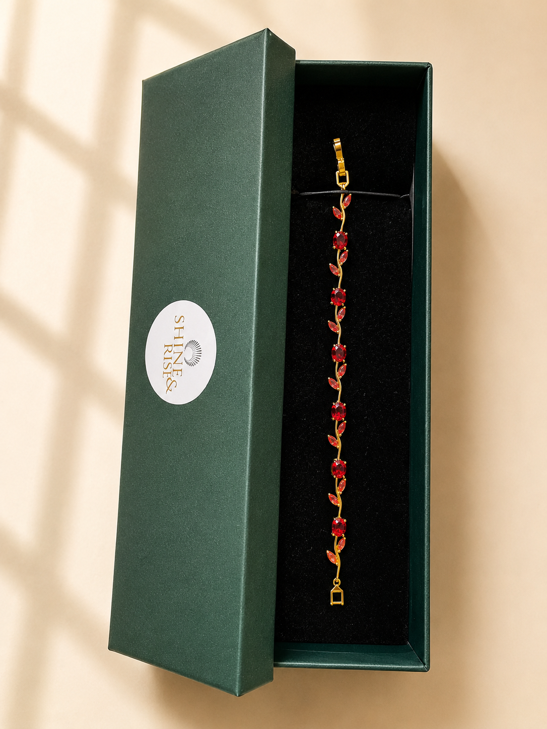 A gold-tone bracelet with red floral links displayed inside an open dark green jewelry box with a black velvet interior. The box lid is propped beside it, showing a white circular logo that reads 'SHI RAKSH'.